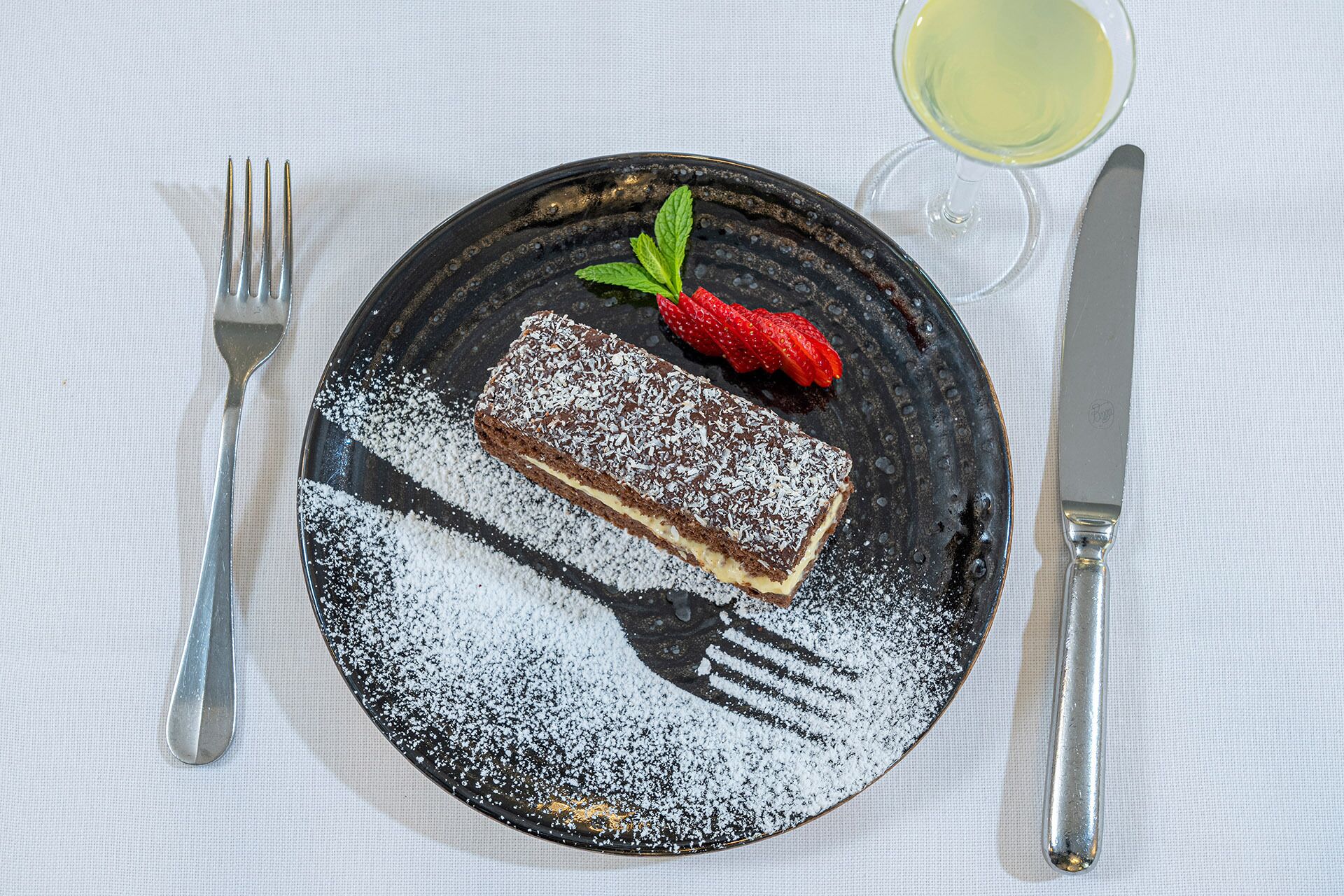 A dessert plate with a piece of cake, a strawberry, and a glass of white wine on a tablecloth.