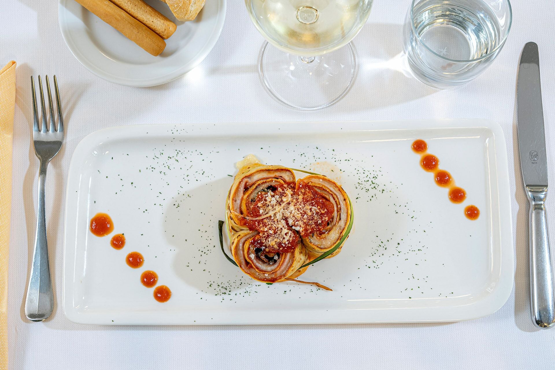 A rectangular white plate with a rolled pastry, garnished with herbs and drizzled with sauce, accompanied by a fork and knife on a white tablecloth.