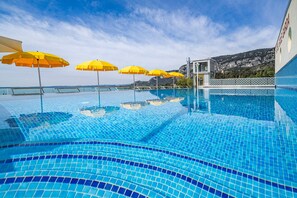 A swimming pool with clear blue water, surrounded by yellow umbrellas, and a view of the sea and hills in the background. - Sky Pool Hotel Sole Garda (Garda)
