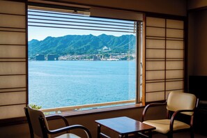 A room with a large window offering a view of a body of water and mountains. There are two chairs and a small table. - Hotel Miyarikyu (Hatsukaichi)