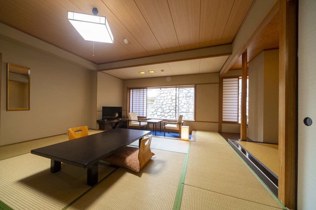 A traditional Japanese room with tatami flooring, a low table, chairs, a television, and a stone wall.