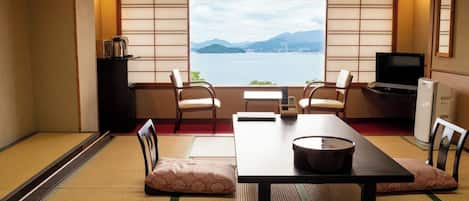 Una habitación japonesa tradicional con suelo de tatami, una mesa baja, sillas, televisión y vistas a un lago con montañas.