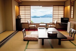 Una habitación japonesa tradicional con suelo de tatami, una mesa baja, sillas, televisión y vistas a un lago con montañas.