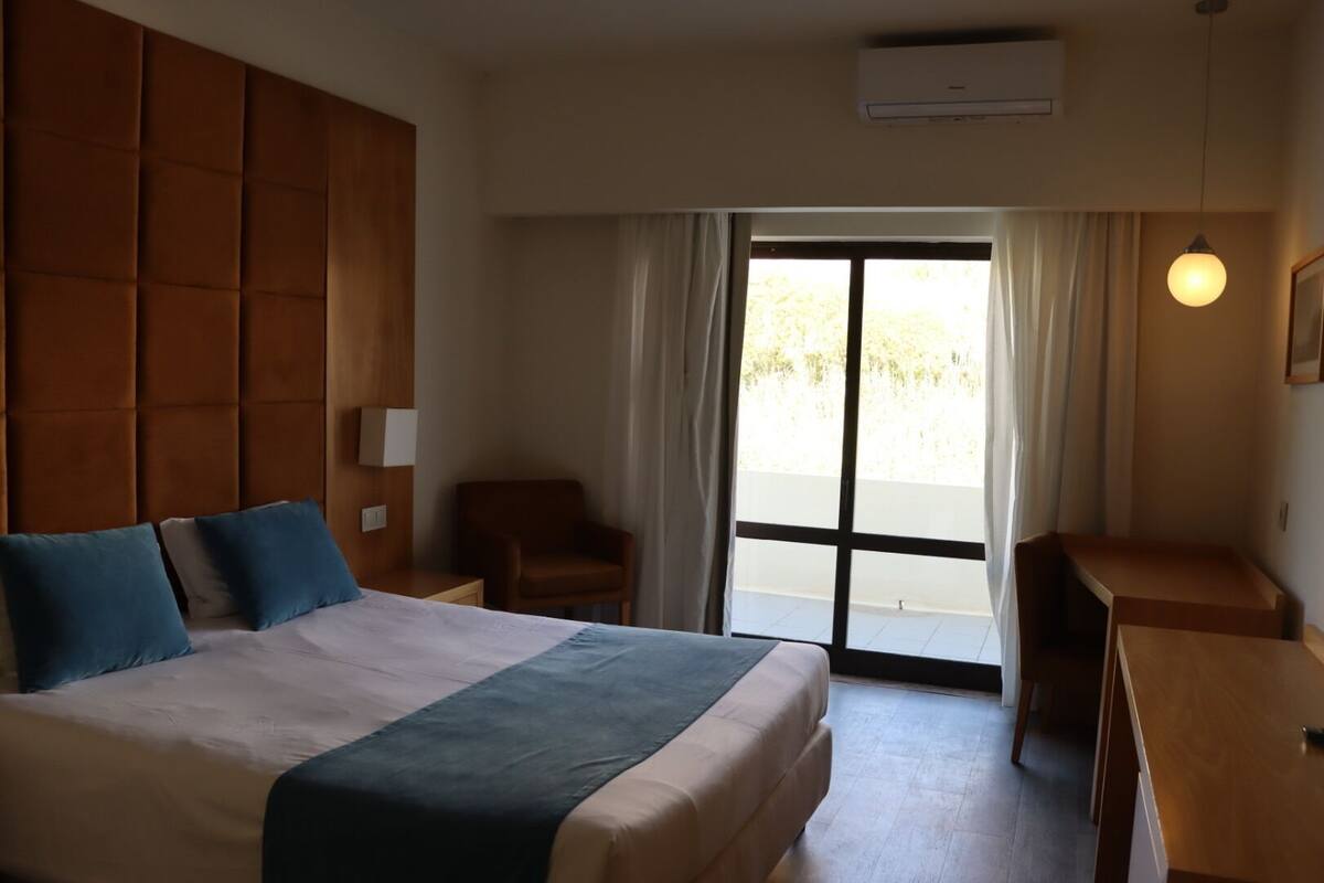 A hotel room with a large bed, a desk, a chair, and a view through a sliding door.