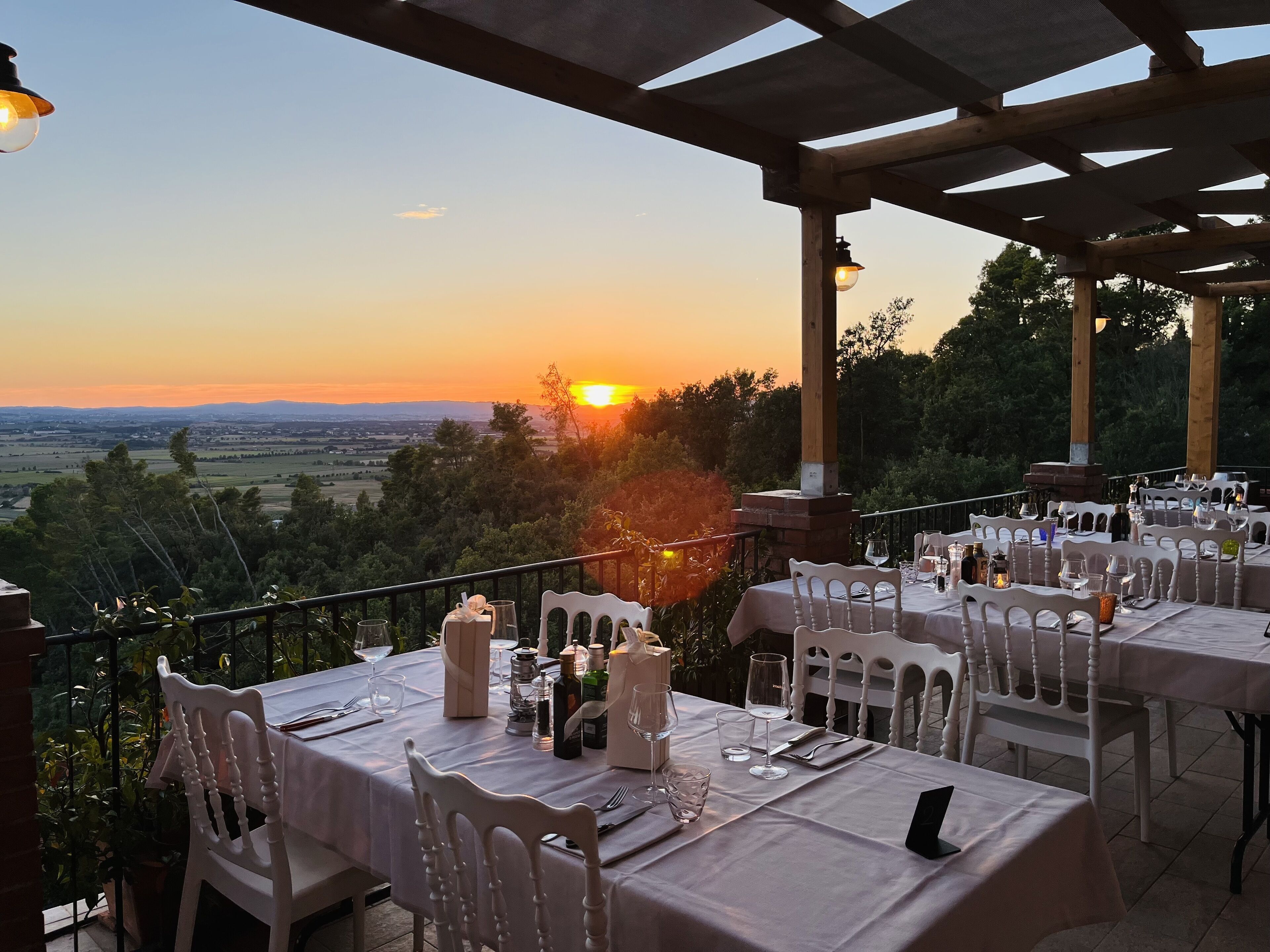 An outdoor dining area with tables set for two, overlooking a scenic landscape during sunset.