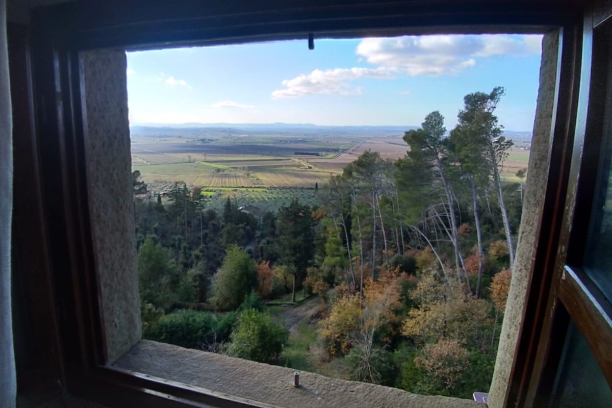 Une vue depuis une fenêtre donnant sur une vallée boisée, avec des champs et des montagnes qui se dessinent à l’horizon.