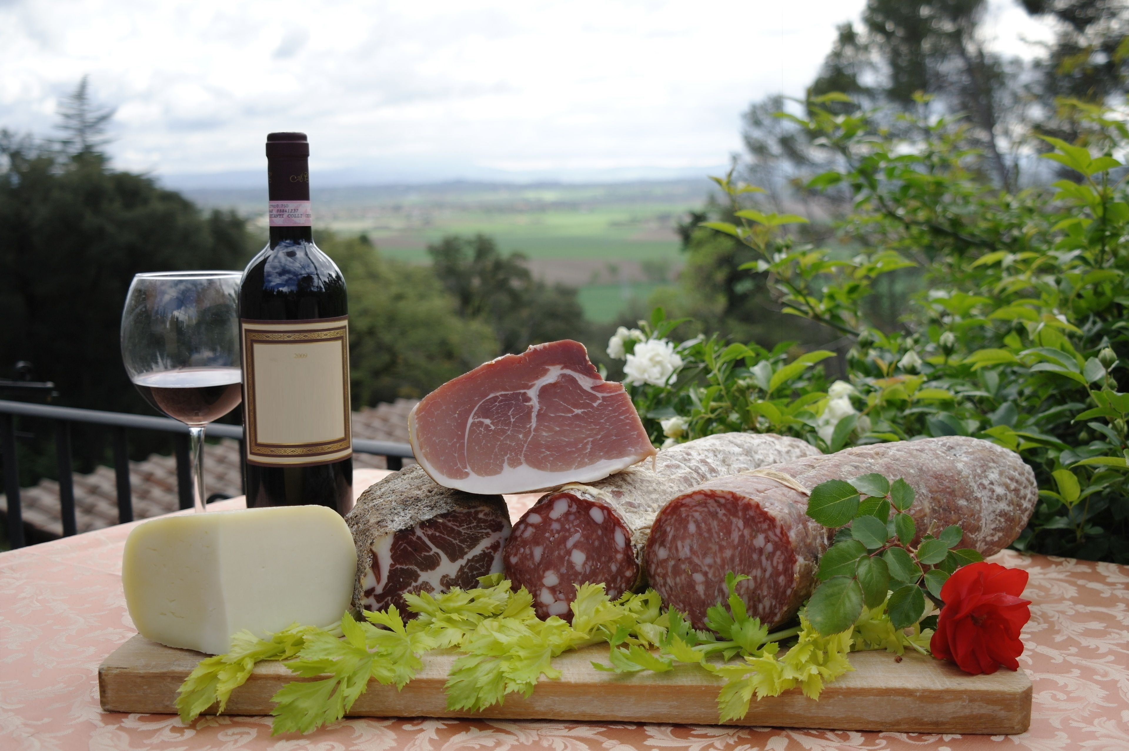 Une table avec une bouteille de vin, un verre, divers fromages, de la charcuterie et une rose rouge.