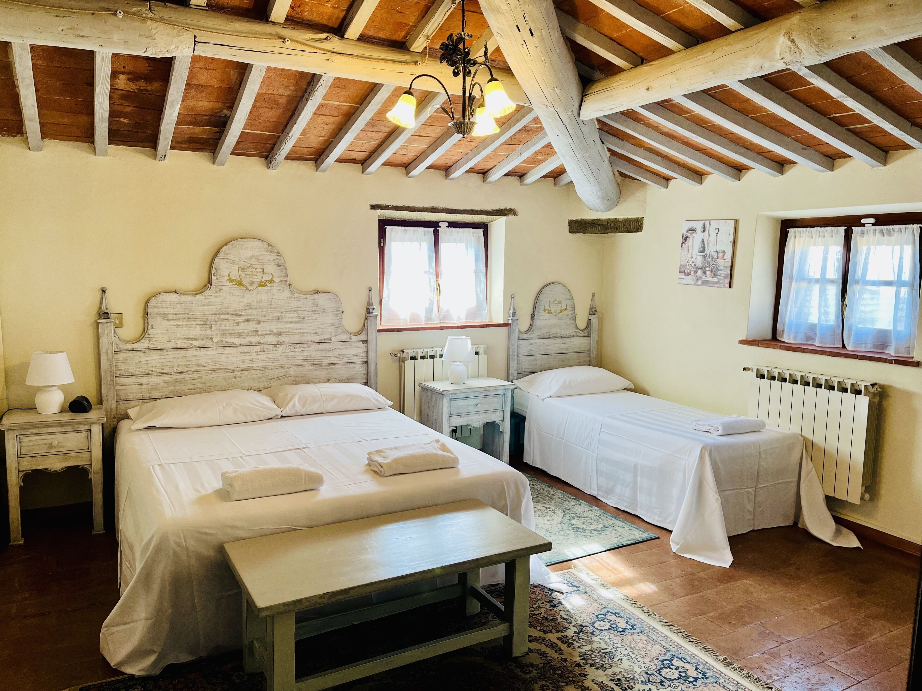 A room with two beds, a bench, a nightstand, a lamp, and a window with curtains.