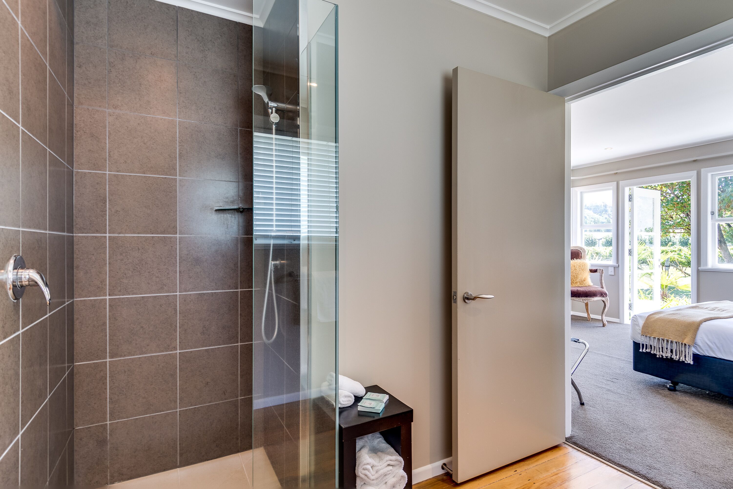 A modern bathroom with a glass-enclosed shower, a wooden bench, and a door leading to a bedroom with a bed and a view of greenery.