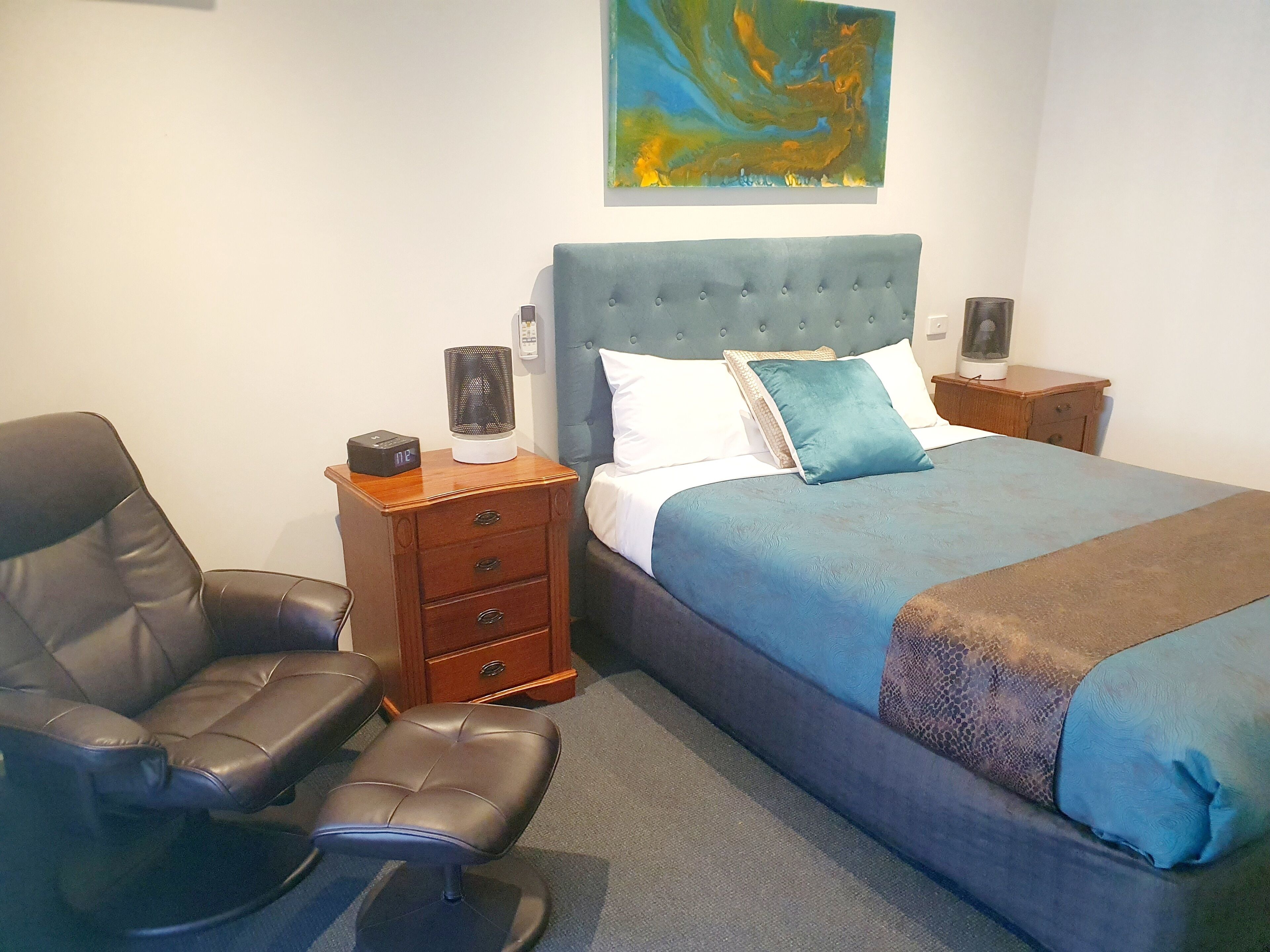 A hotel room with a bed, a chair, a nightstand, and a painting on the wall.