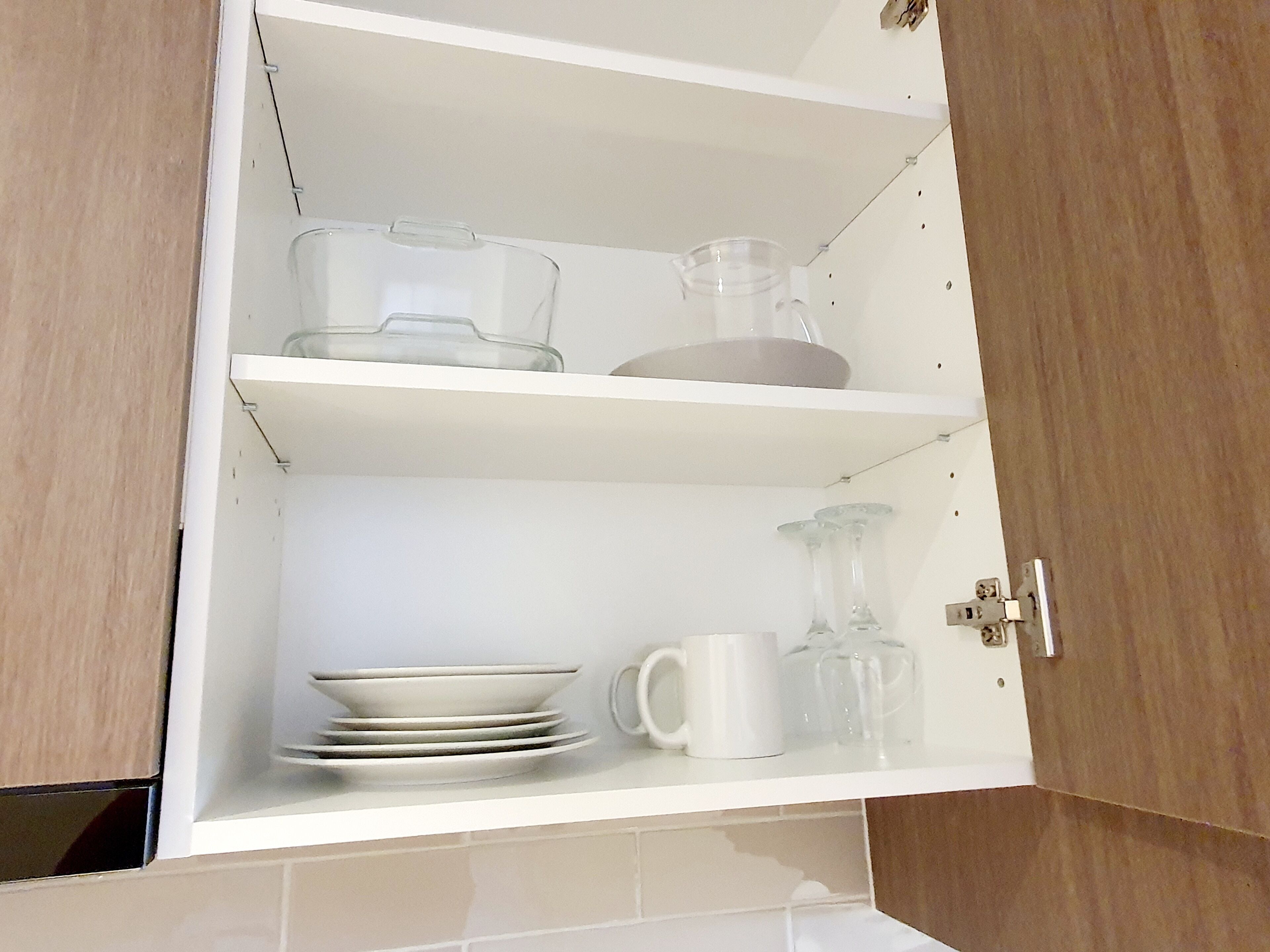 Een keukenkastje met planken waarop drinkglazen, borden en een kan staan.