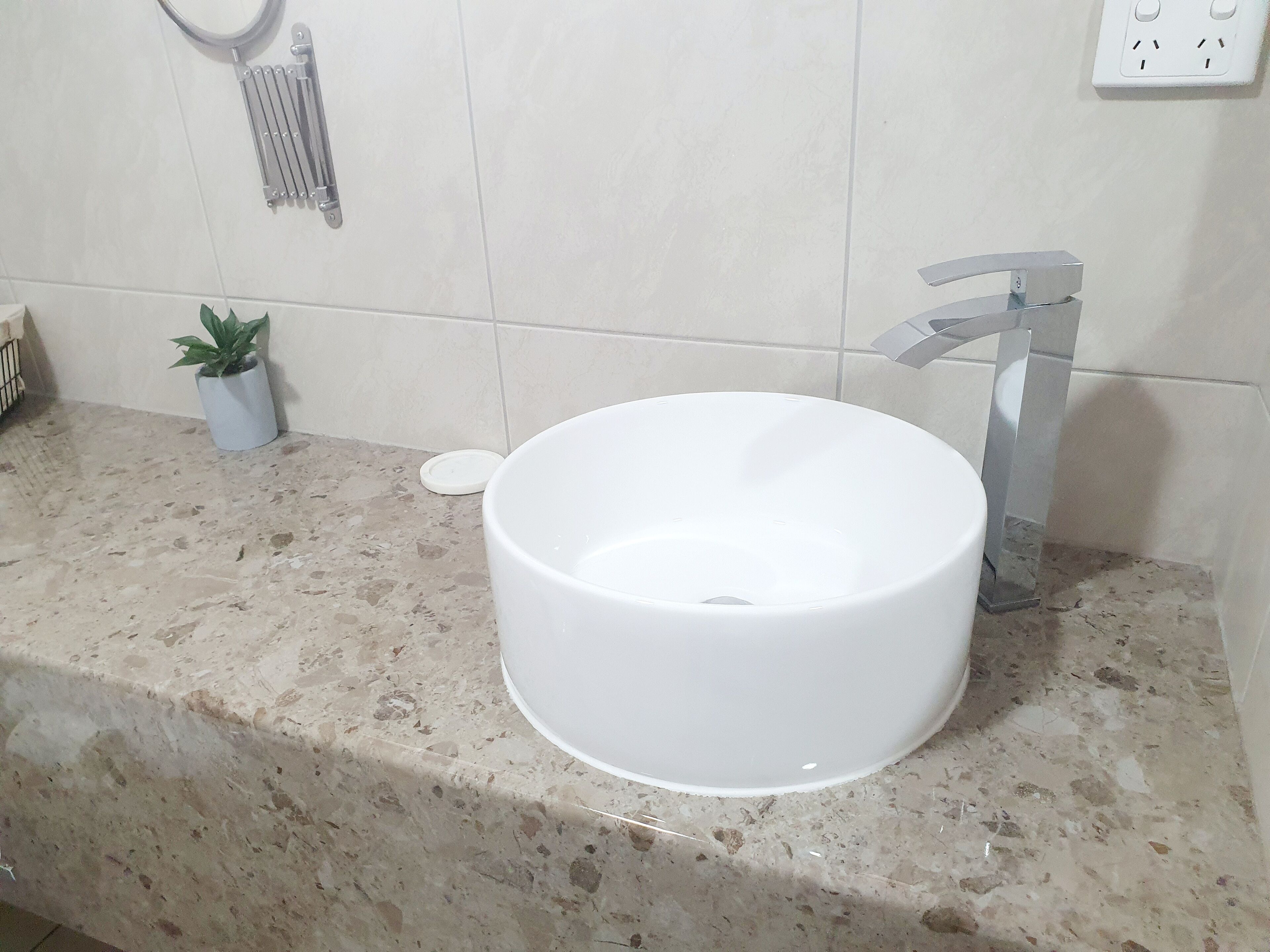 Un baño con un lavamanos blanco, una pequeña planta en maceta, un perchero y un enchufe en la pared.