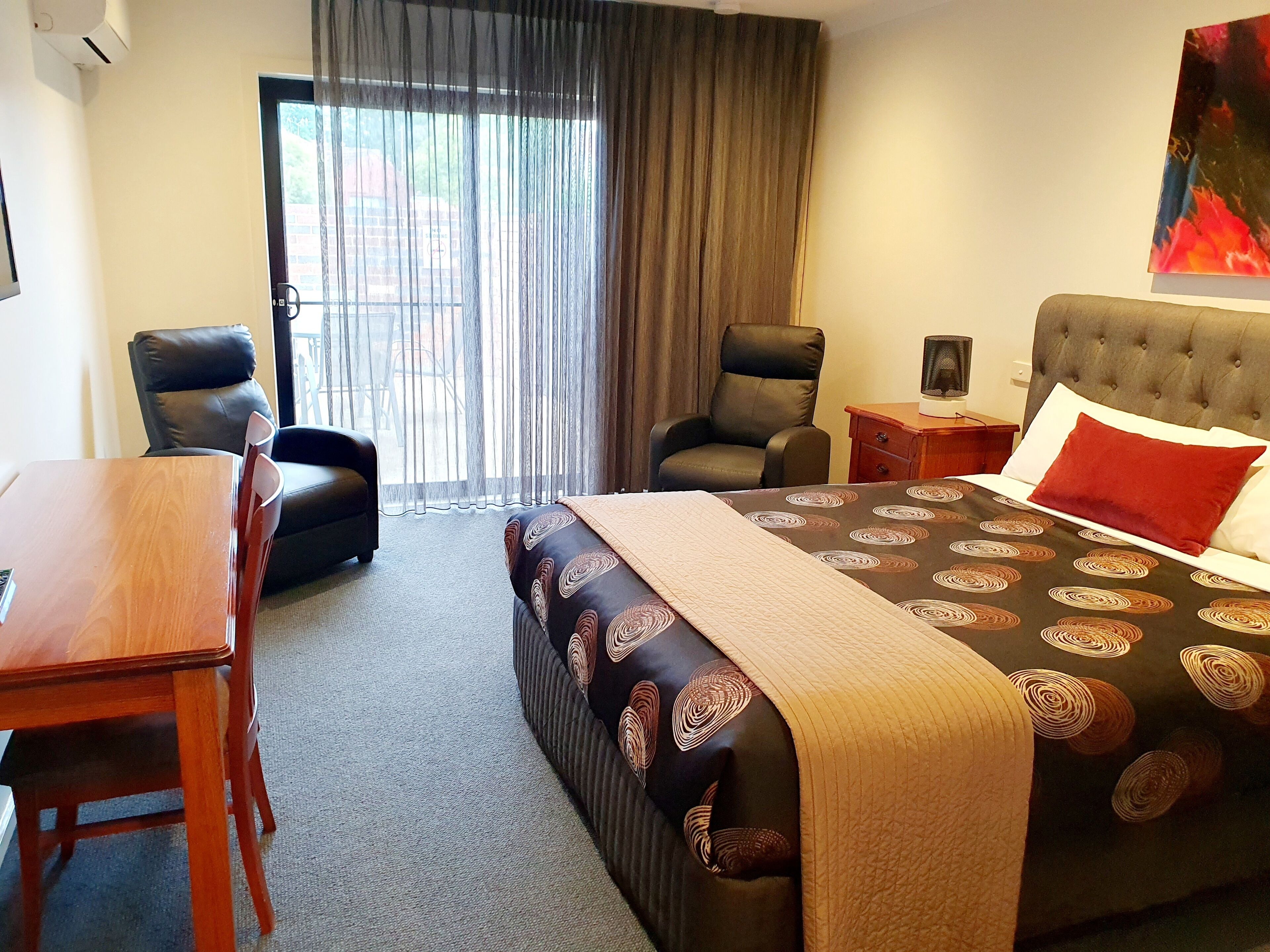 A hotel room with a bed, two armchairs, a wooden table, a TV, and a large window with curtains.