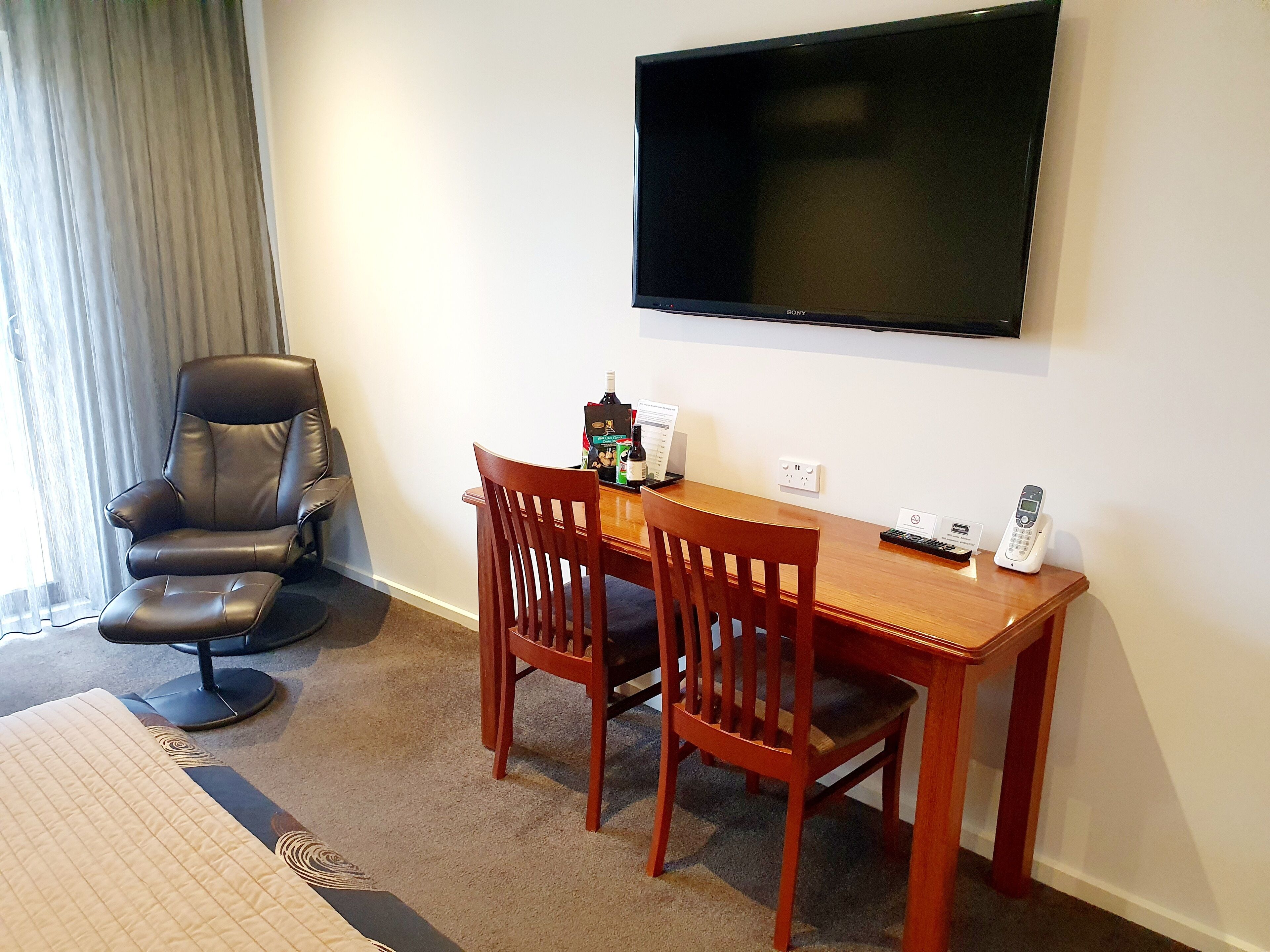 Ein Hotelzimmer mit einem Holztisch, zwei Stühlen, einem Ledersessel, einem flachbildfernseher an der Wand, einem Telefon und einem kleinen Tisch mit einer Flasche und einer Speisekarte.