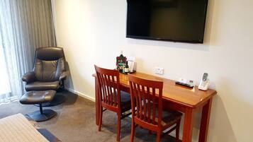 Ein Hotelzimmer mit einem Holztisch, zwei Stühlen, einem Ledersessel, einem flachbildfernseher an der Wand, einem Telefon und einem kleinen Tisch mit einer Flasche und einer Speisekarte.