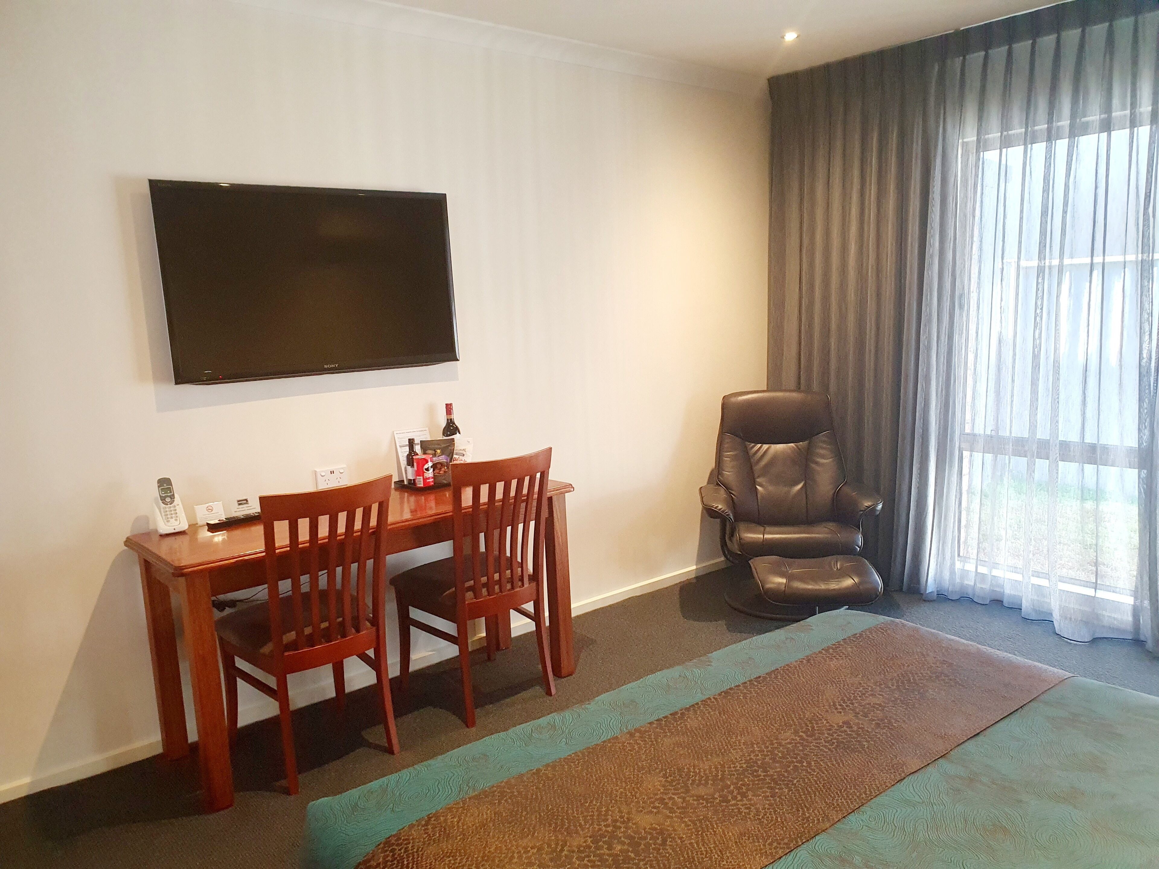 A hotel room with a flat-screen TV mounted on the wall, a wooden dining table with two chairs, a leather armchair, and a large window with sheer curtains.
