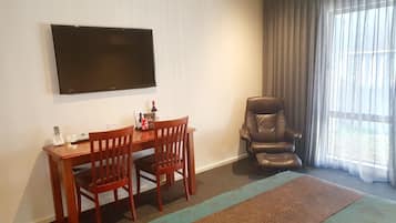 A hotel room with a flat-screen TV mounted on the wall, a wooden dining table with two chairs, a leather armchair, and a large window with sheer curtains.