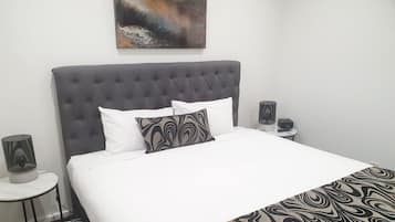 Cama bem arrumada com cabeceira cinza, almofadas brancas e estampadas, e manta decorativa. Tem uma mesinha de cabeceira com luminária e um objeto decorativo.