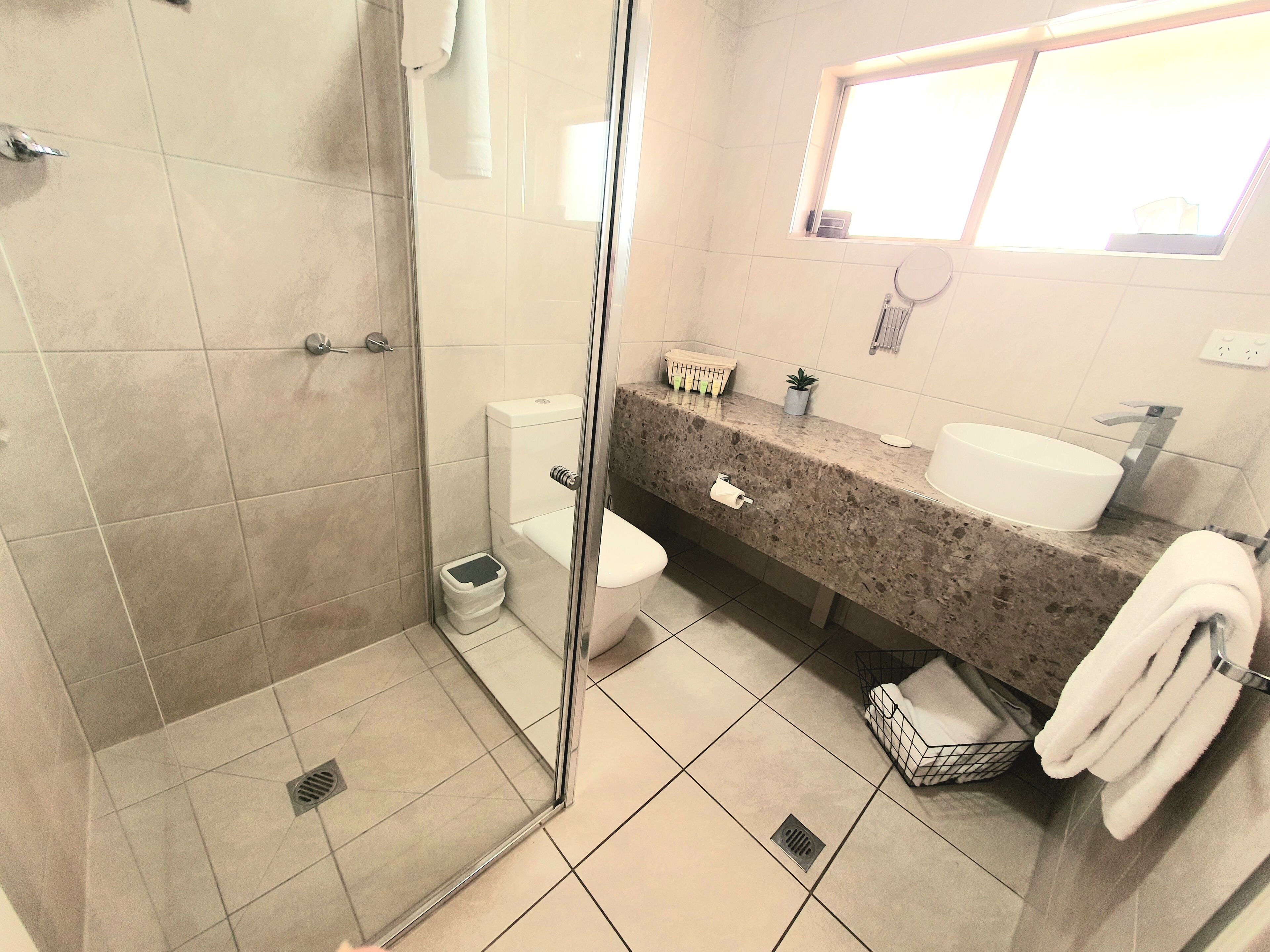 Een moderne badkamer met een glazen douchecabine, een toilet, een wastafel met een marmeren aanrechtblad en een klein raam boven de wastafel.