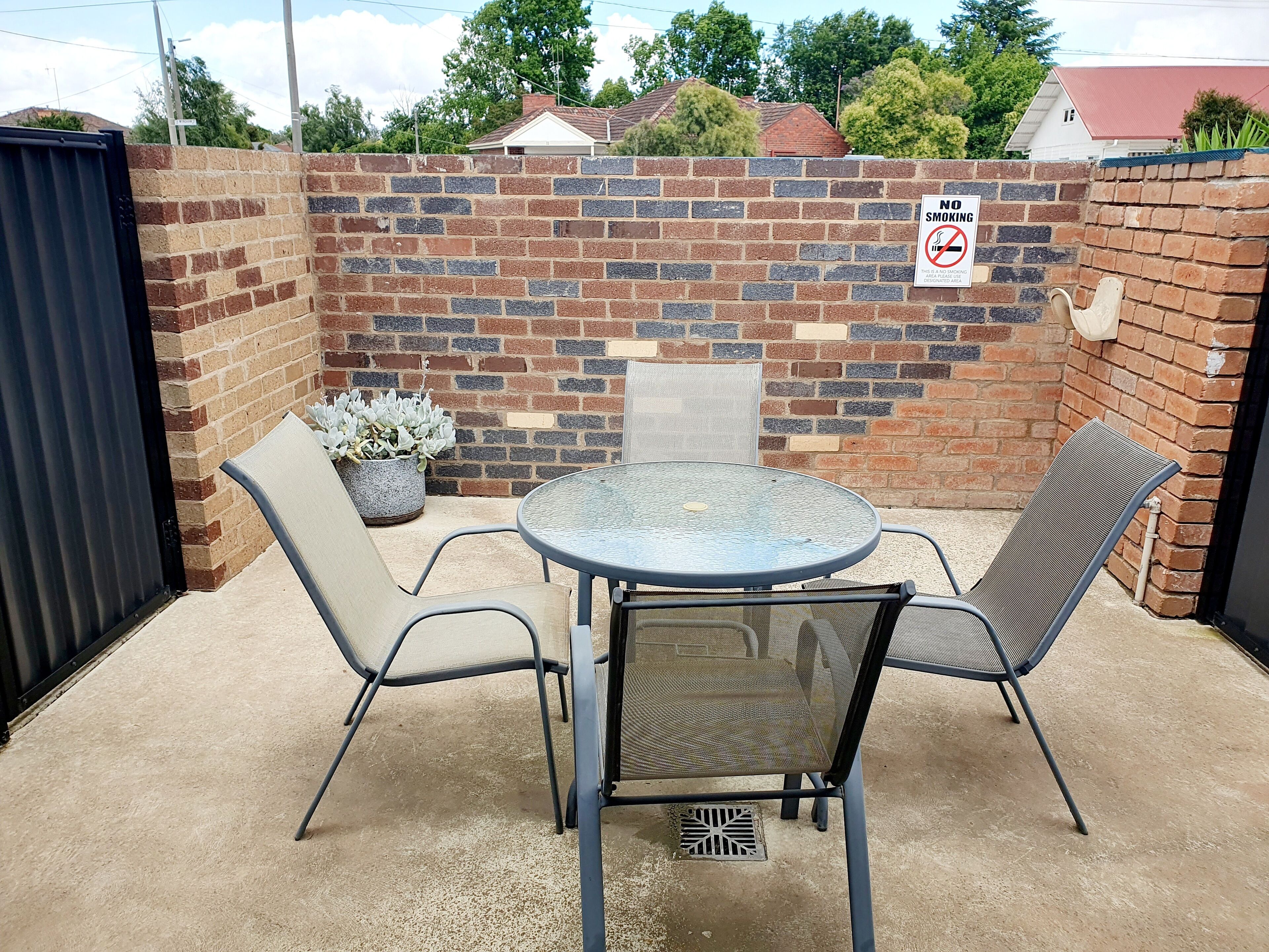A patio area with a round metal table and two chairs, a potted plant, and a brick wall with a 'No Smoking' sign.