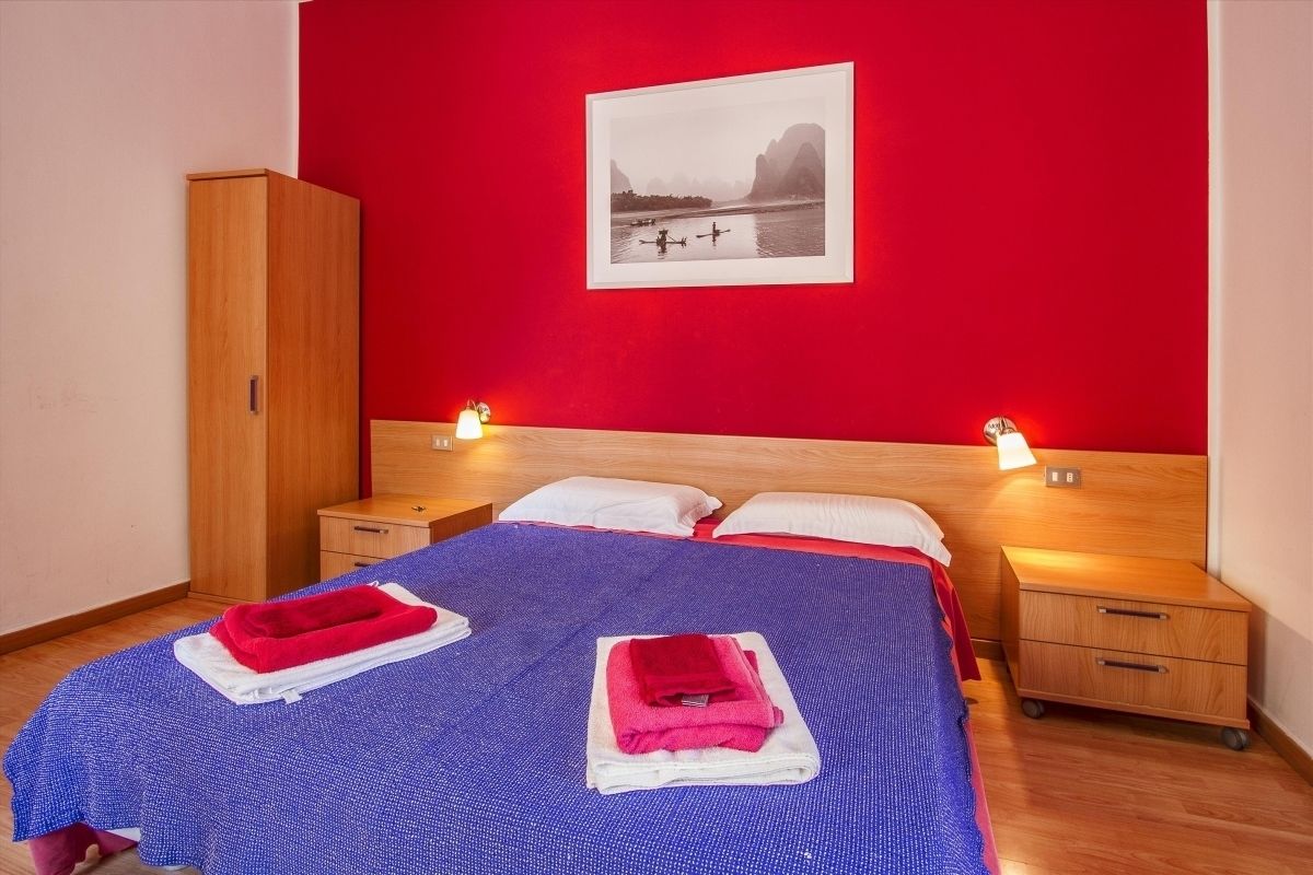 A hotel room with a red wall, a bed with blue bedding, two white pillows, a wooden wardrobe, and a framed picture of a landscape.