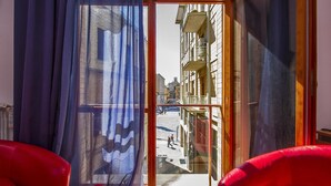 Habitación con un gran ventanal que da a un espacio exterior, con una silla roja y vista a un edificio con balcones.
