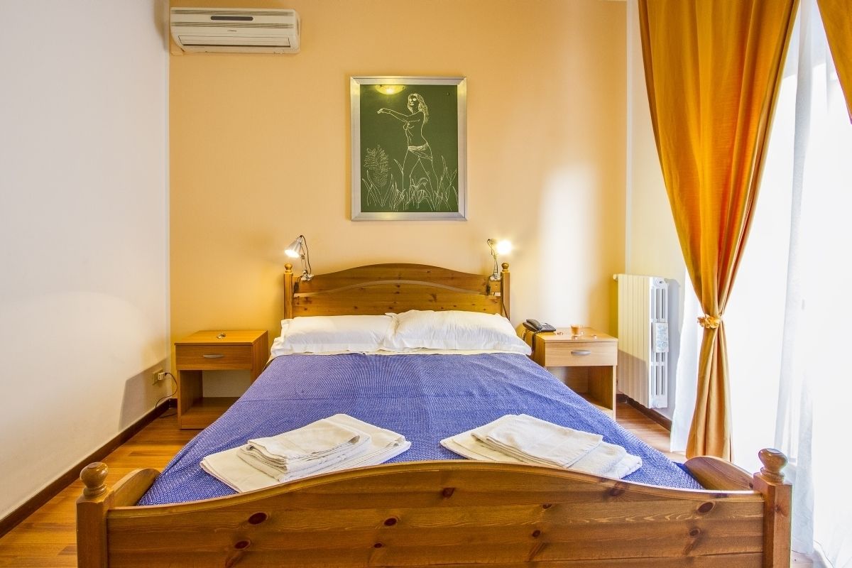 Una cama bien hecha con una colcha azul y almohadas blancas, una mesita de noche con una lámpara y un cuadro en la pared.