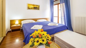 Habitación de hotel con piso de madera, cama con ropa de cama azul, mesita de noche con lámpara y un jarrón con girasoles.