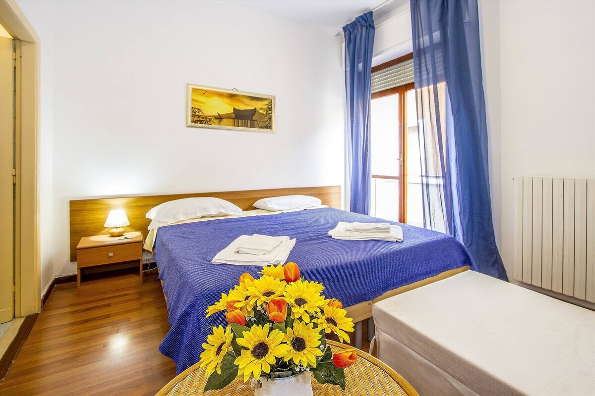 Ein Hotelzimmer mit Holzboden, einem Bett mit blauer Bettwäsche, einem Nachttisch mit Lampe und einer Vase mit Sonnenblumen.