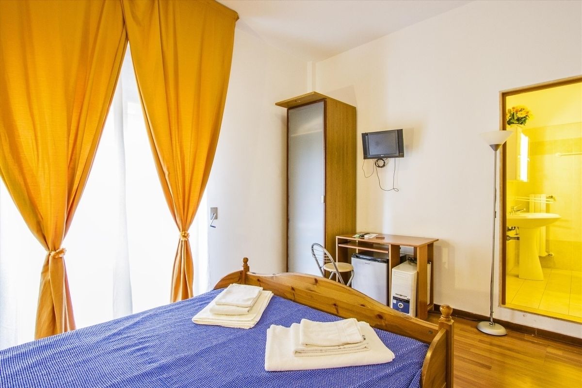 A bedroom with a bed, a desk, a chair, a television, and a bathroom with a yellow sink.