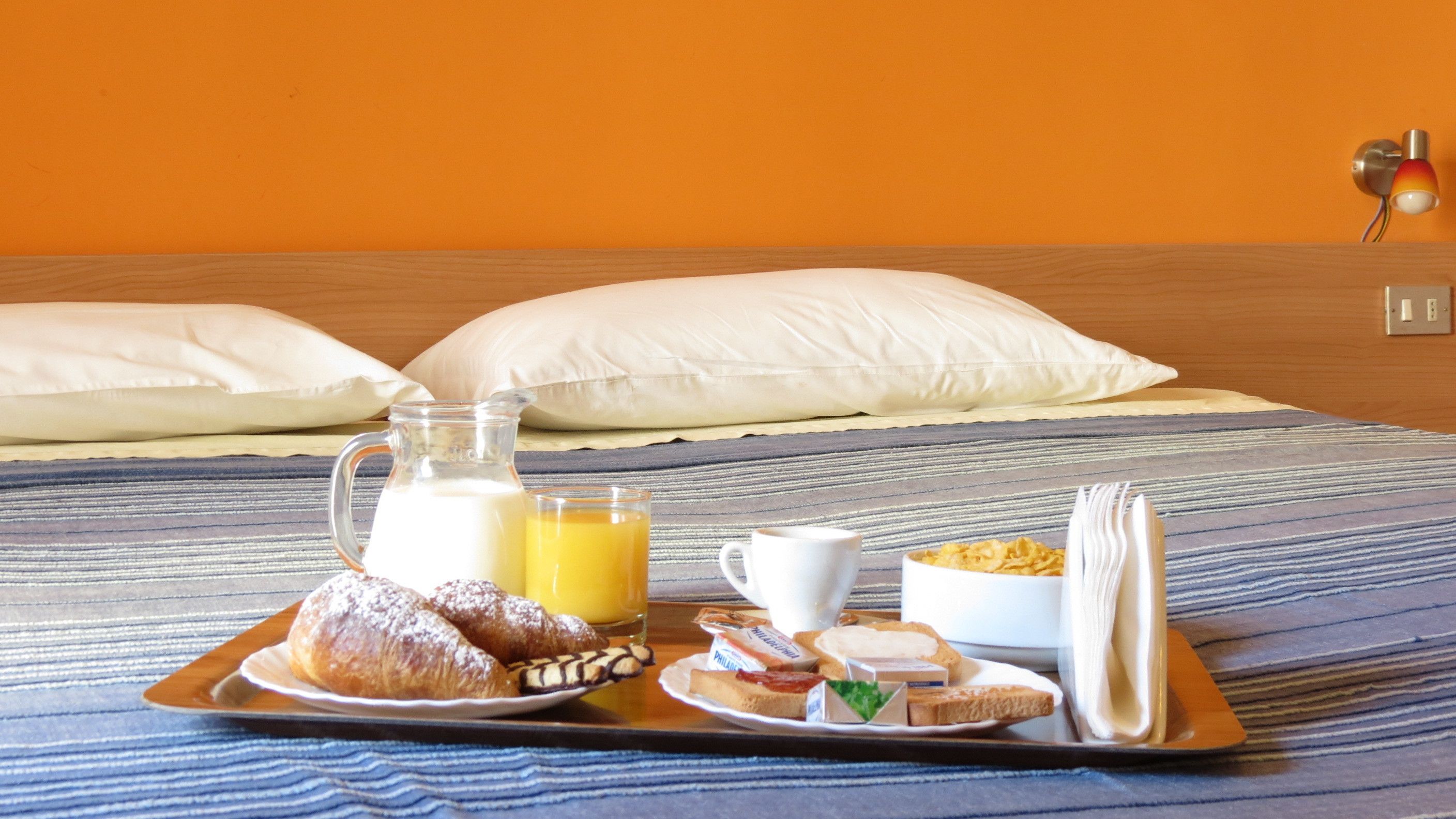 En bakke med morgenmad på sengen, med et stribet tæppe, inklusive croissanter, appelsinjuice, mælk, cornflakes og toast, samt en kop.