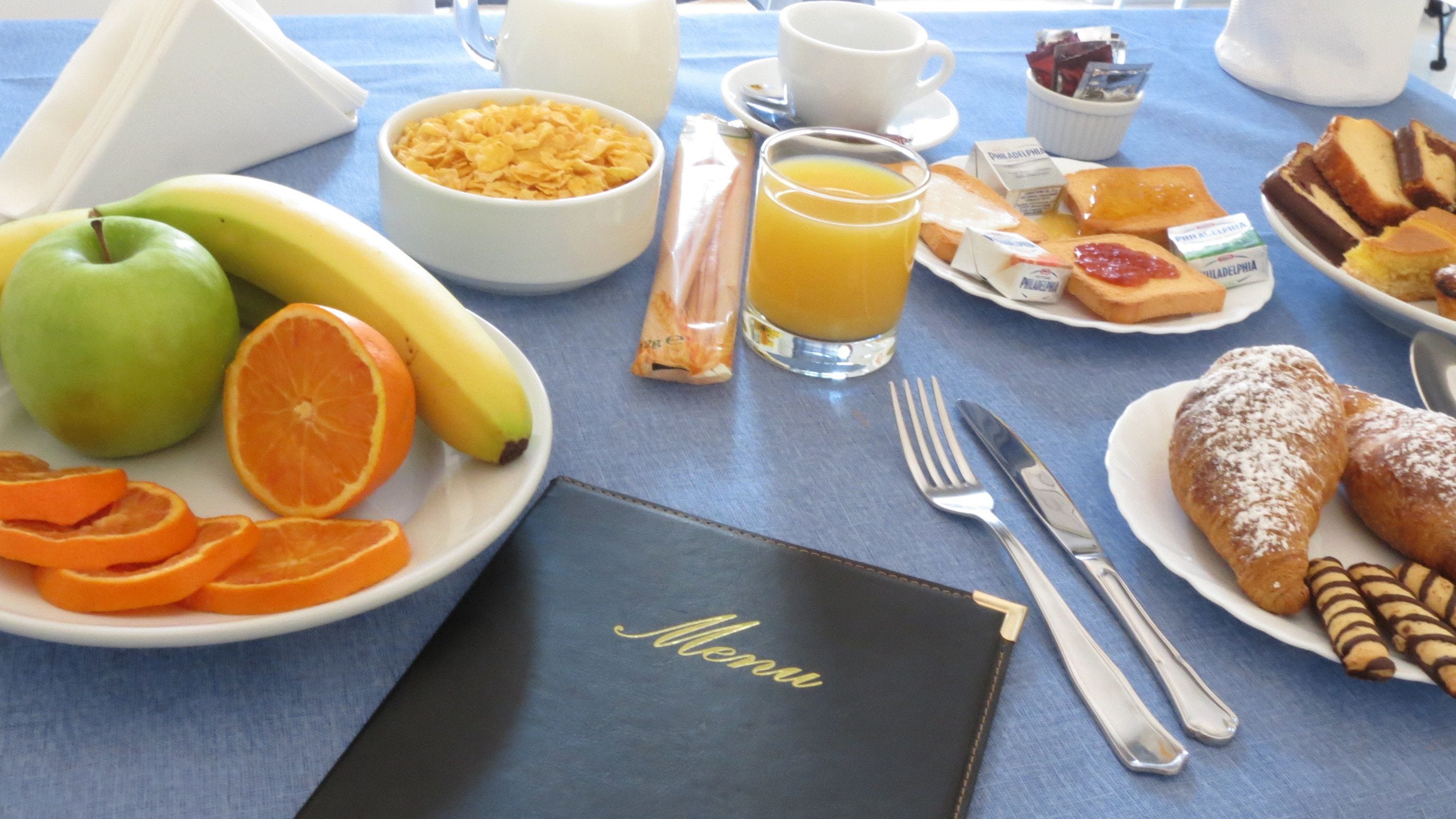 A breakfast table with a menu, fruits, cereal, orange juice, and pastries.