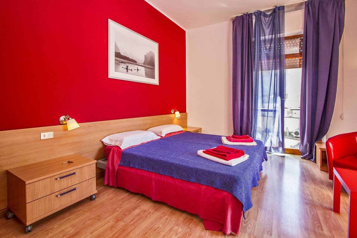 Ein Hotelzimmer mit roter Wand, einem Bett mit blauer und roter Bettdecke, einem Nachttisch aus Holz, einer Holzkommode und einem Fenster mit violetten Vorhängen.