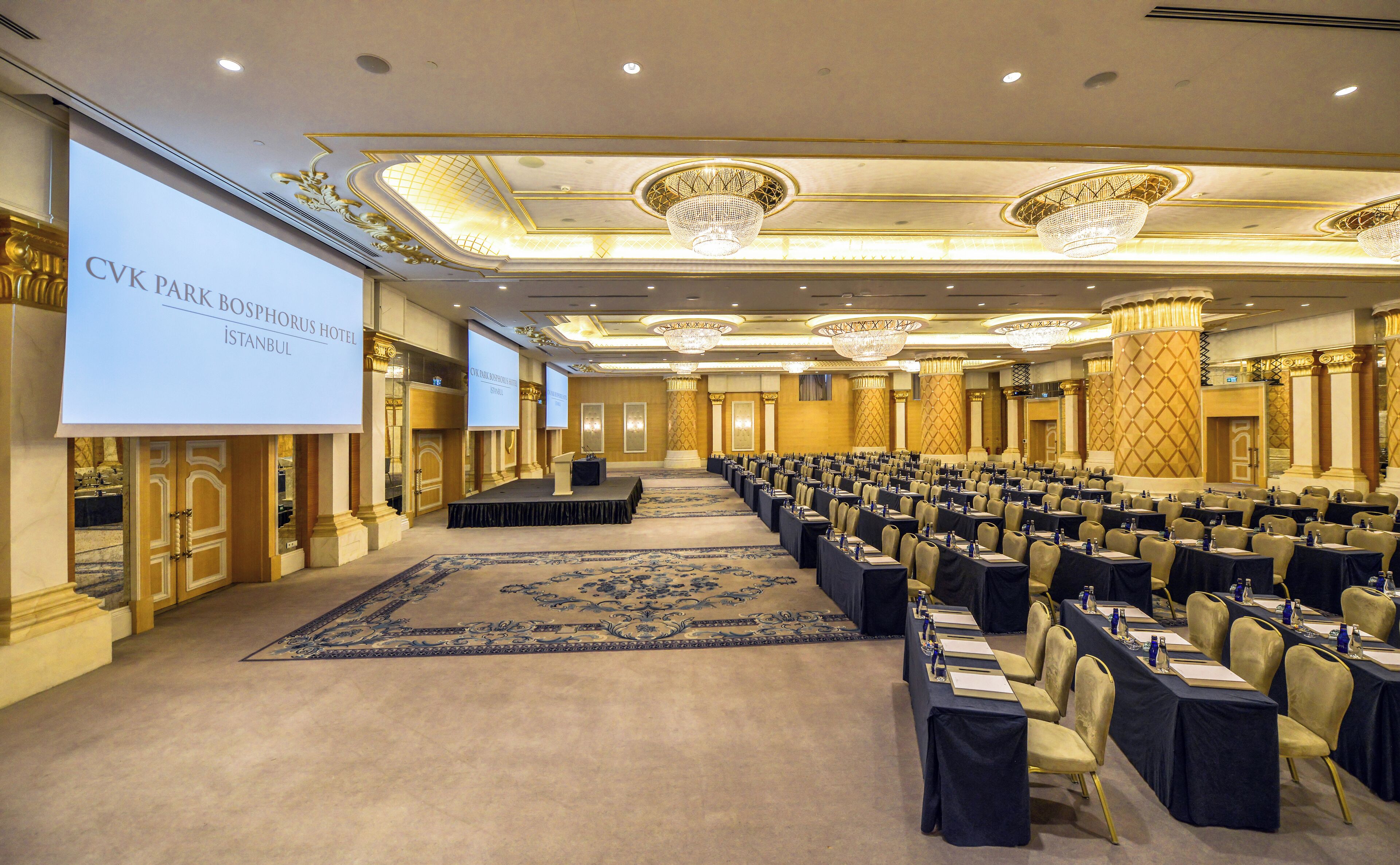 A conference room with rows of tables and chairs, a large screen, and chandeliers.