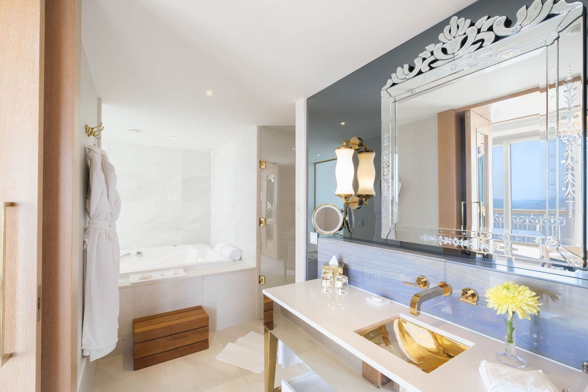 A modern bathroom with a large mirror, gold fixtures, and a view of the sea.