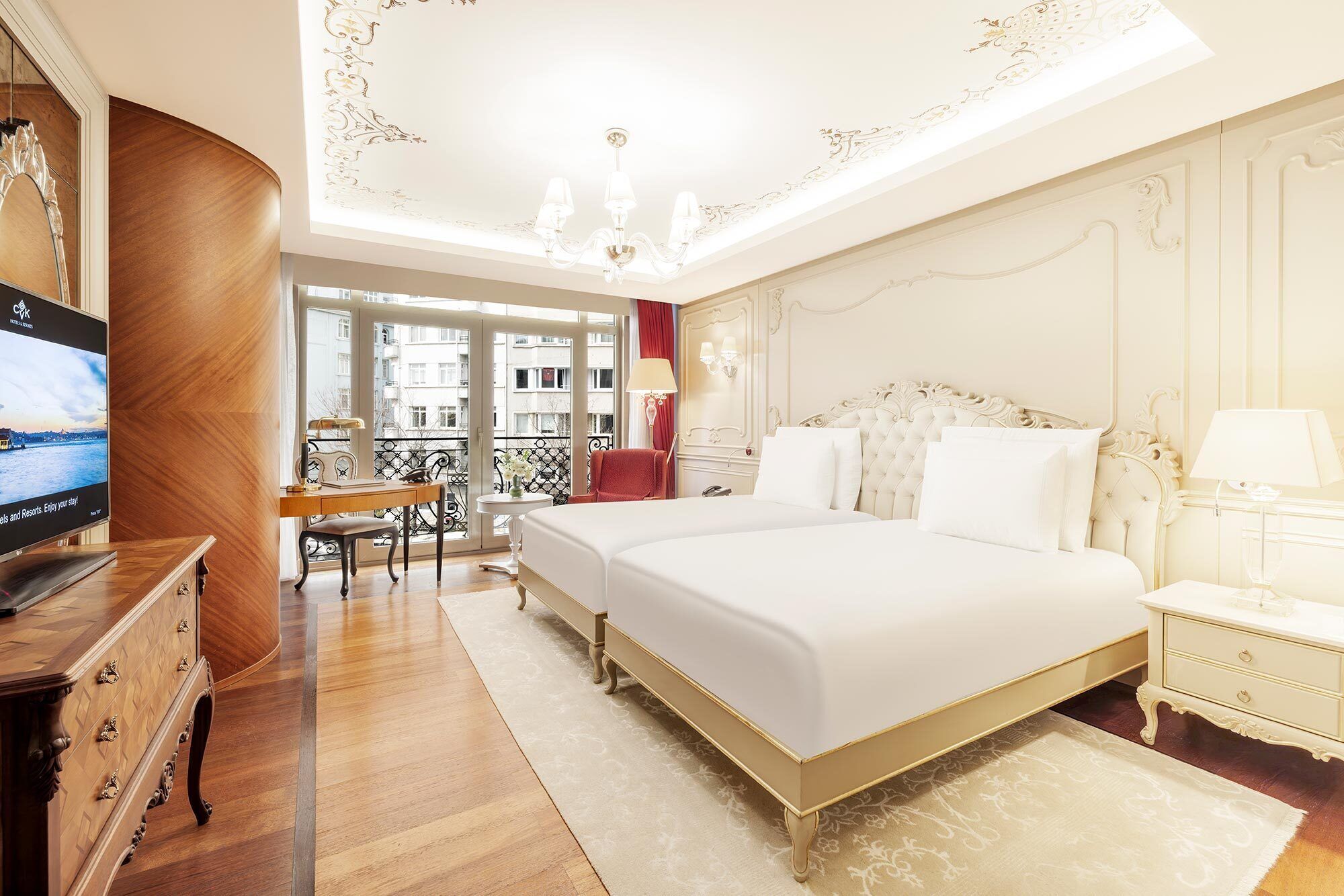 A hotel room with a large bed, a desk with a chair, a television, and a balcony with a view of buildings.