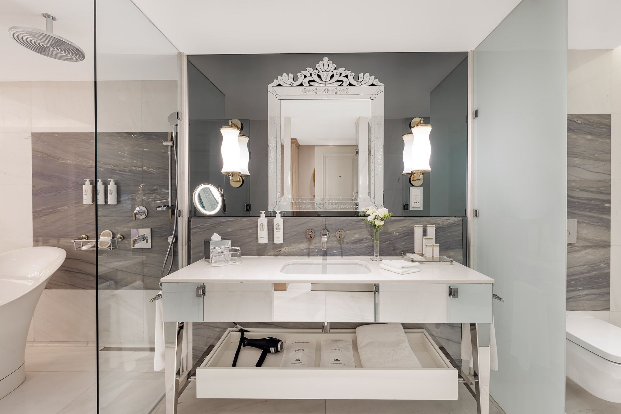 A modern bathroom with a double sink vanity, a large mirror with wall sconces, a glass shower enclosure, and a marble backsplash.