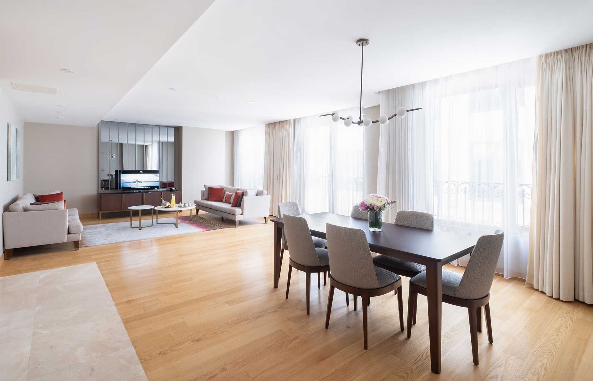 Una sala de estar moderna con comedor, piso de madera y ventanas grandes con cortinas.