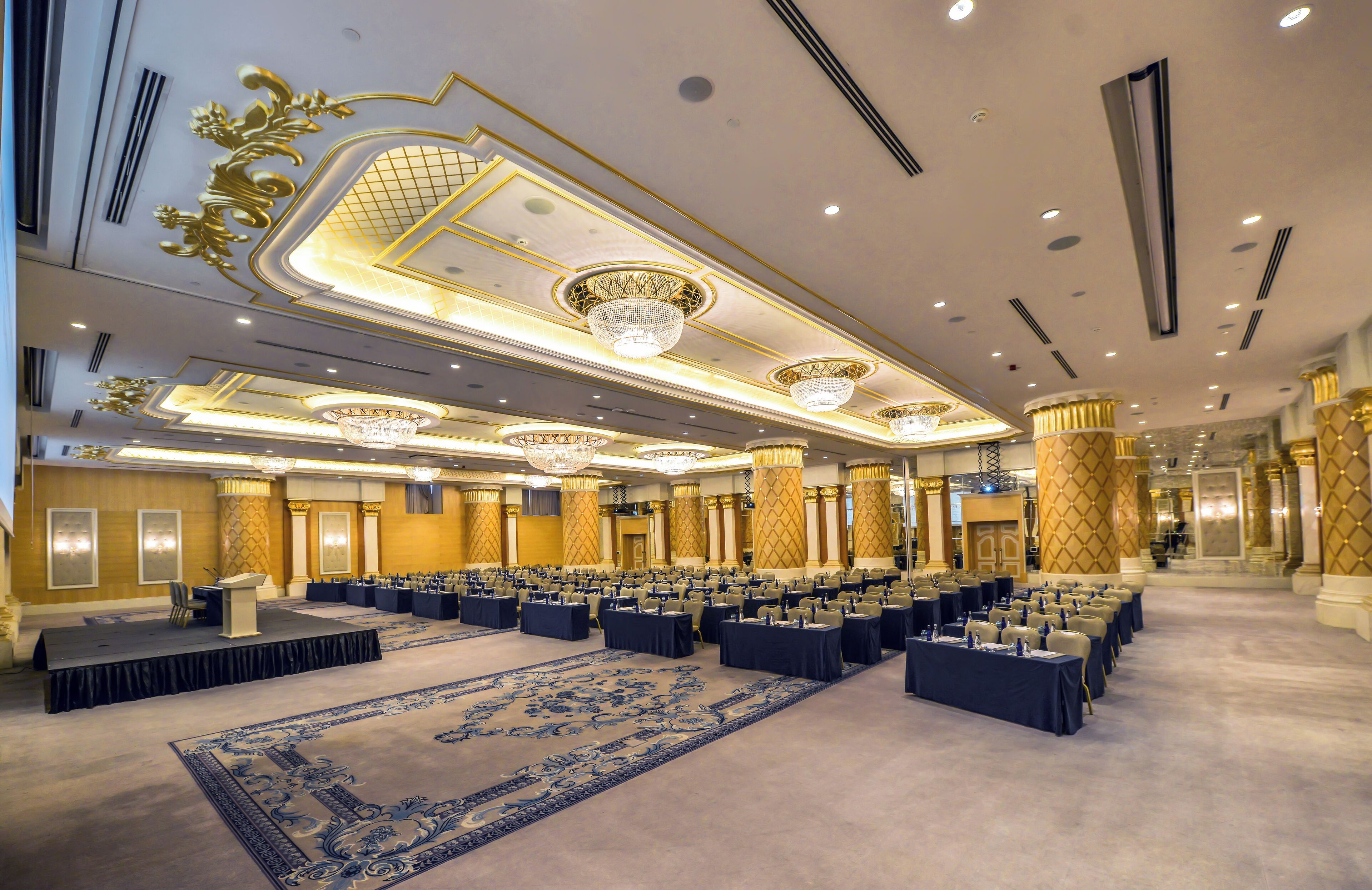 A large conference room with rows of tables and chairs, a stage, and ornate ceiling decorations.