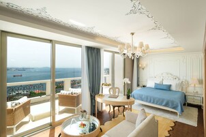 Quarto de hotel luxuoso com uma varanda grande, cama com colcha azul, mesa de jantar redonda e um candelabro.