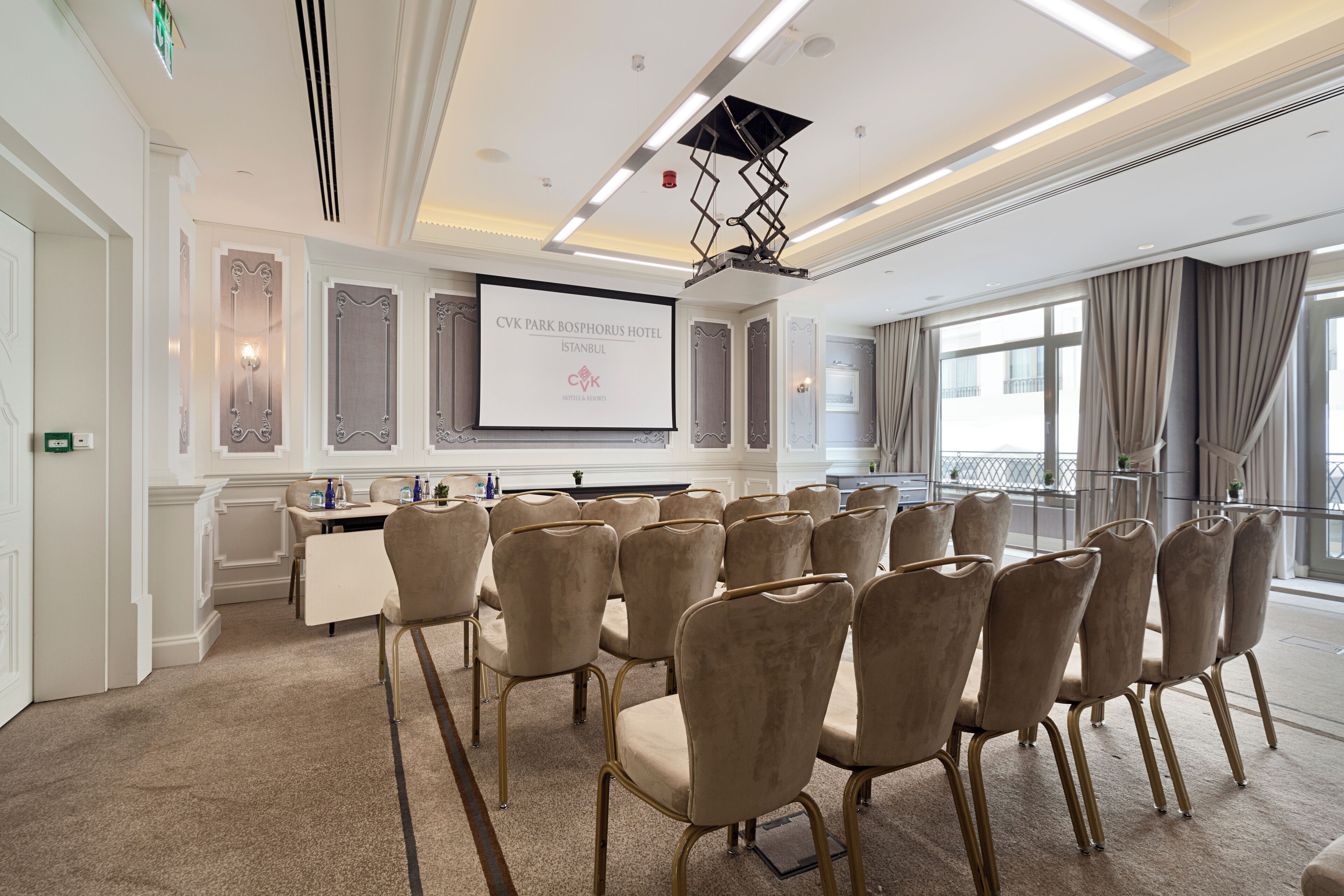 Une salle de conférence avec des chaises de couleur beige, un bureau blanc et un écran de projection affichant le nom de l’hôtel.