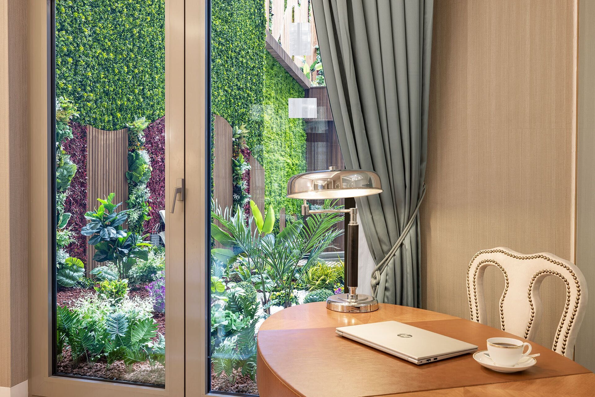 Una habitación con un escritorio de madera, una computadora portátil, una taza de café, una lámpara y vistas a un jardín exuberante a través de una ventana.