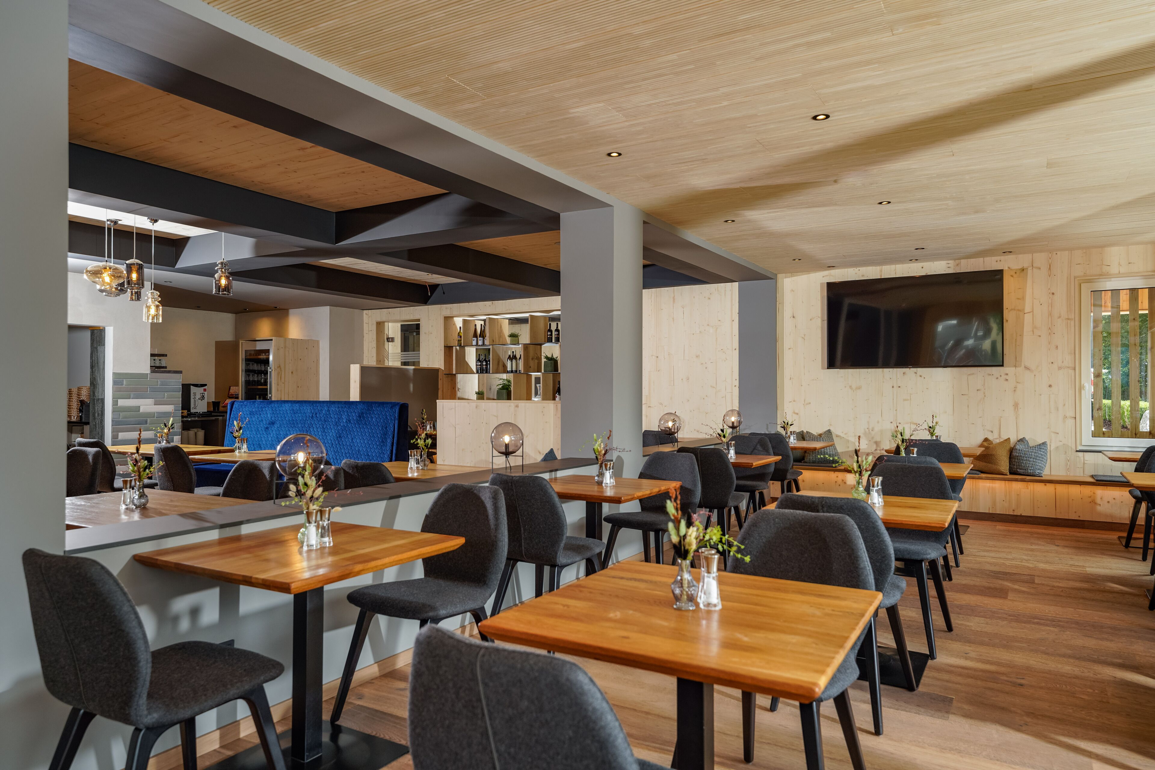 A modern restaurant with wooden tables and chairs, a kitchen area, and a large TV screen on the wall.