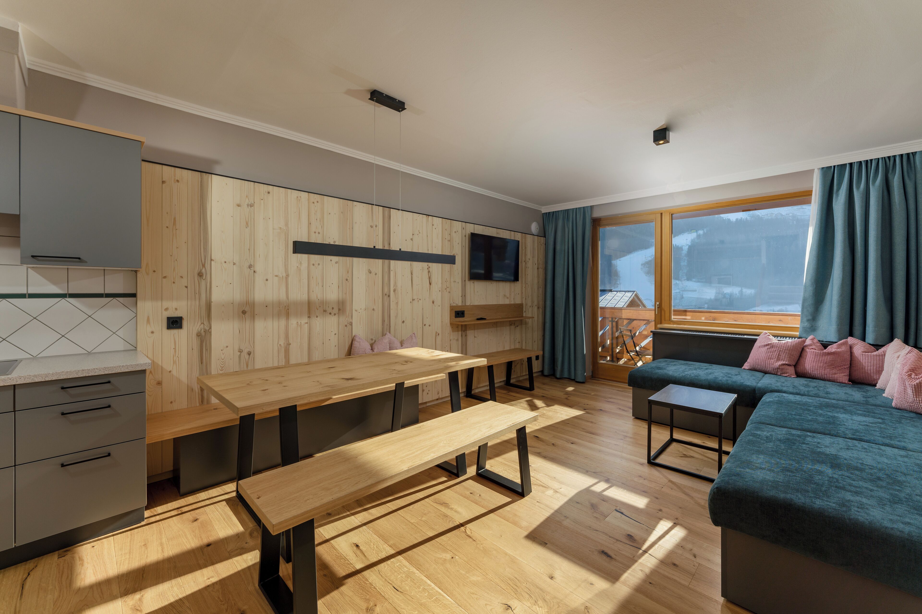 A modern kitchen with wooden paneling, a dining area with benches, a sofa, and a TV mounted on the wall.
