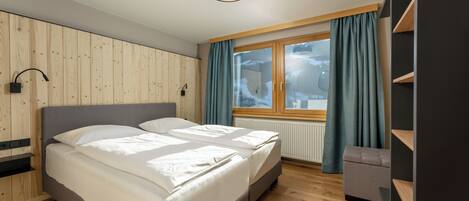 Un dormitorio moderno con un panel de madera, una cama con ropa blanca, un cabecero gris, una ventana con cortinas y una pequeña zona de estar.