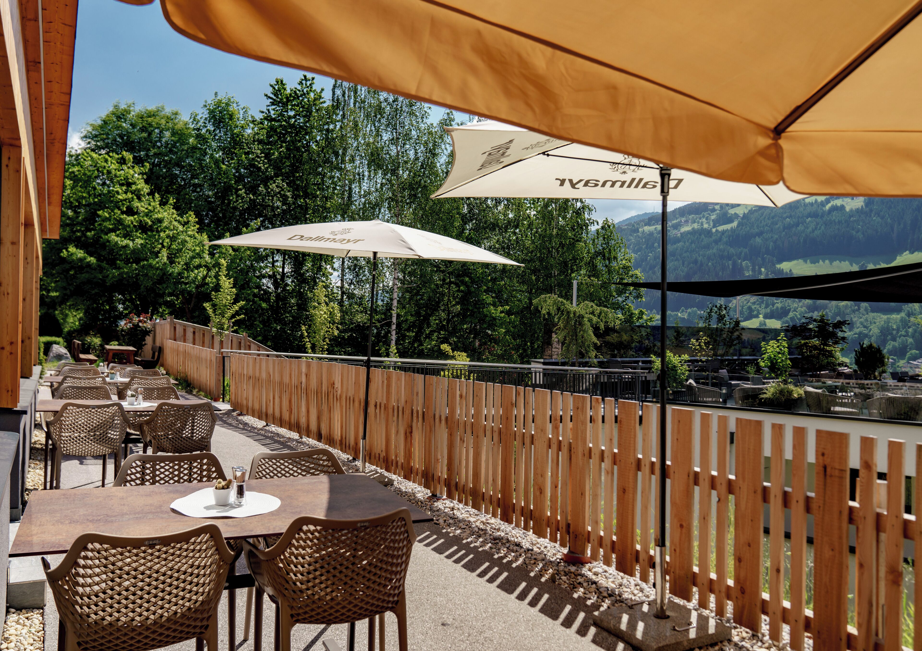 Une terrasse extérieure avec des tables et des chaises, une clôture en bois et des parasols pour créer de l’ombre.