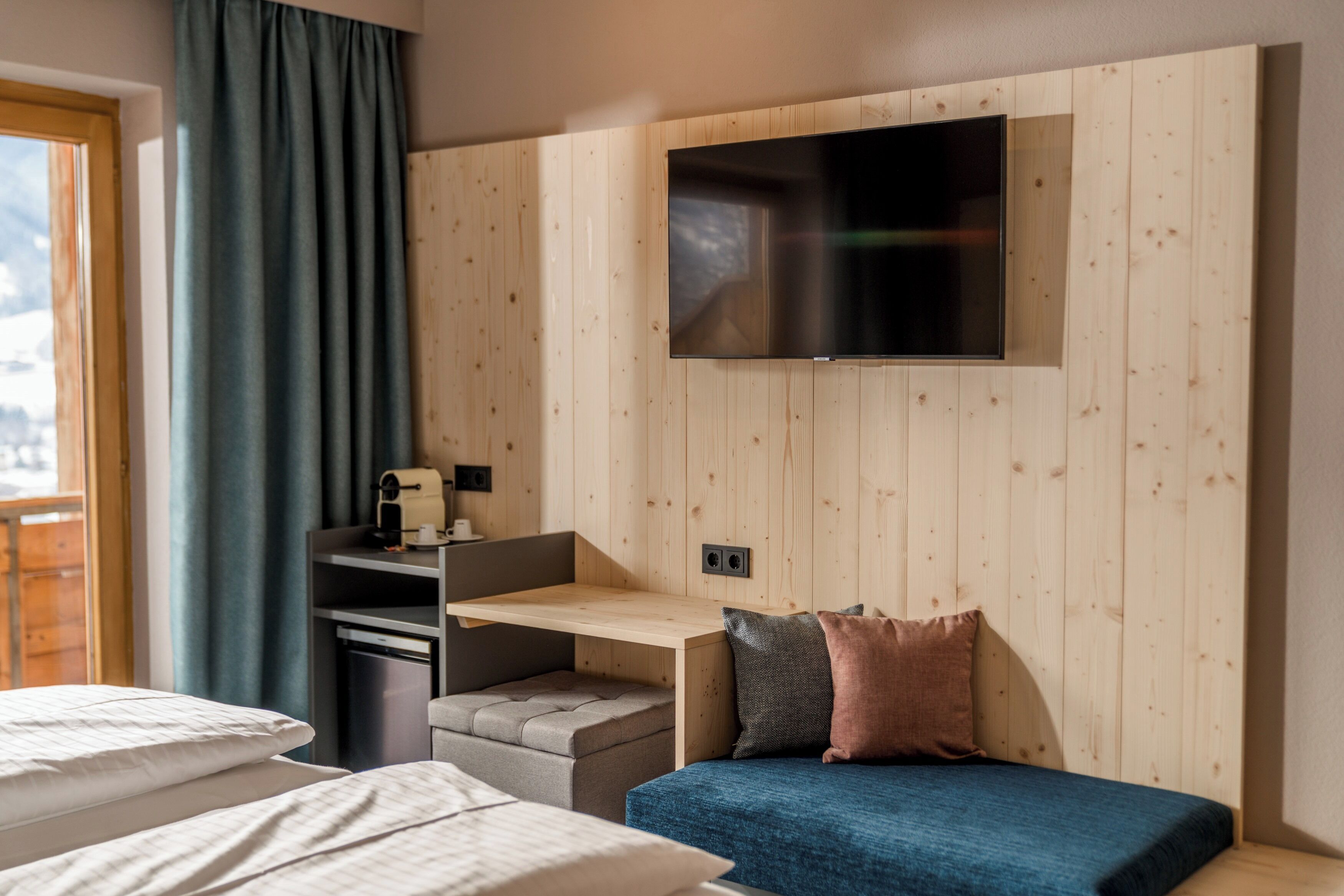 A modern hotel room with a wooden panel wall, a flat-screen TV mounted above a desk, a sofa with cushions, and a window with curtains.