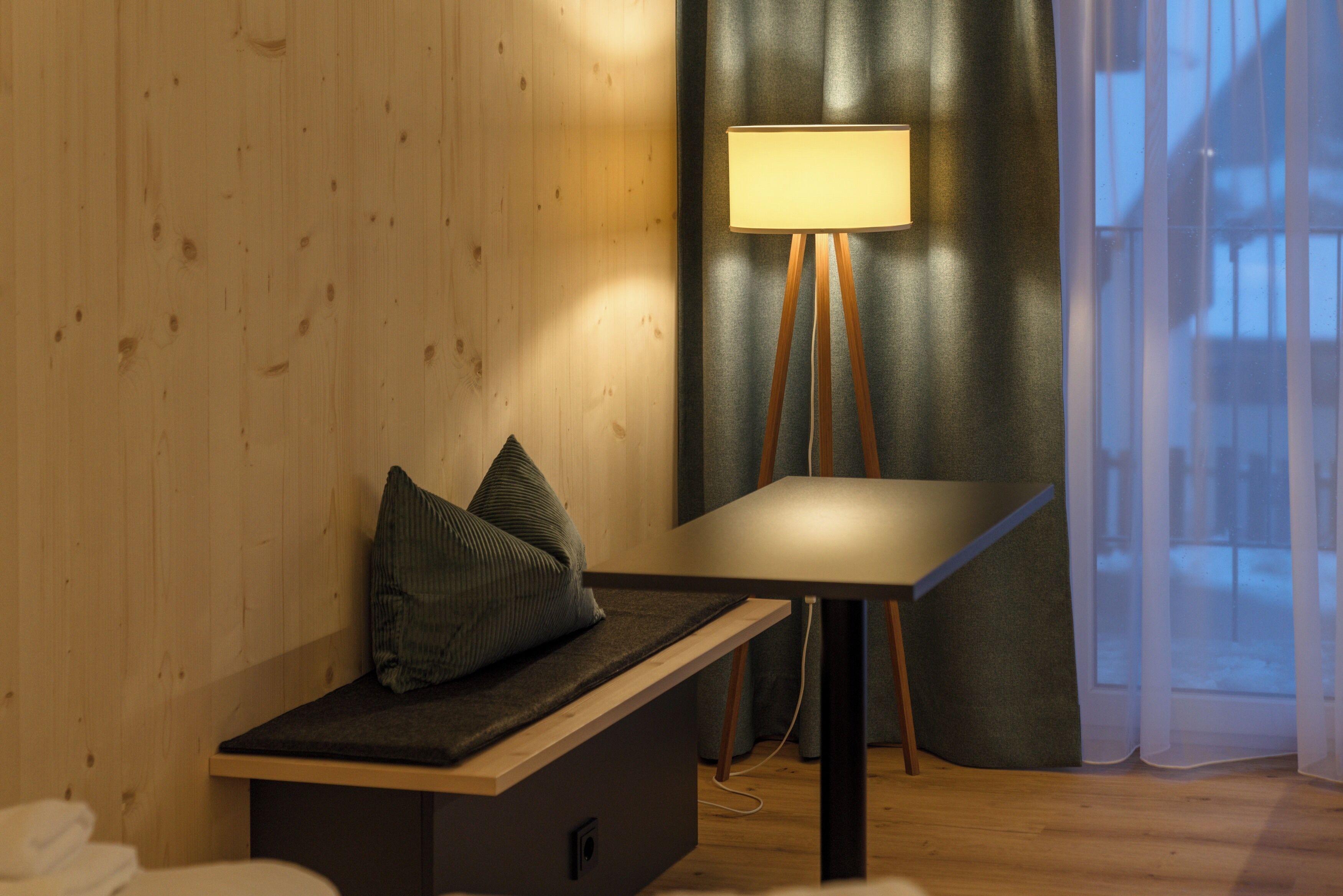 Una habitación con pared de madera, un banco con cojines, una mesa y una lámpara de pie.