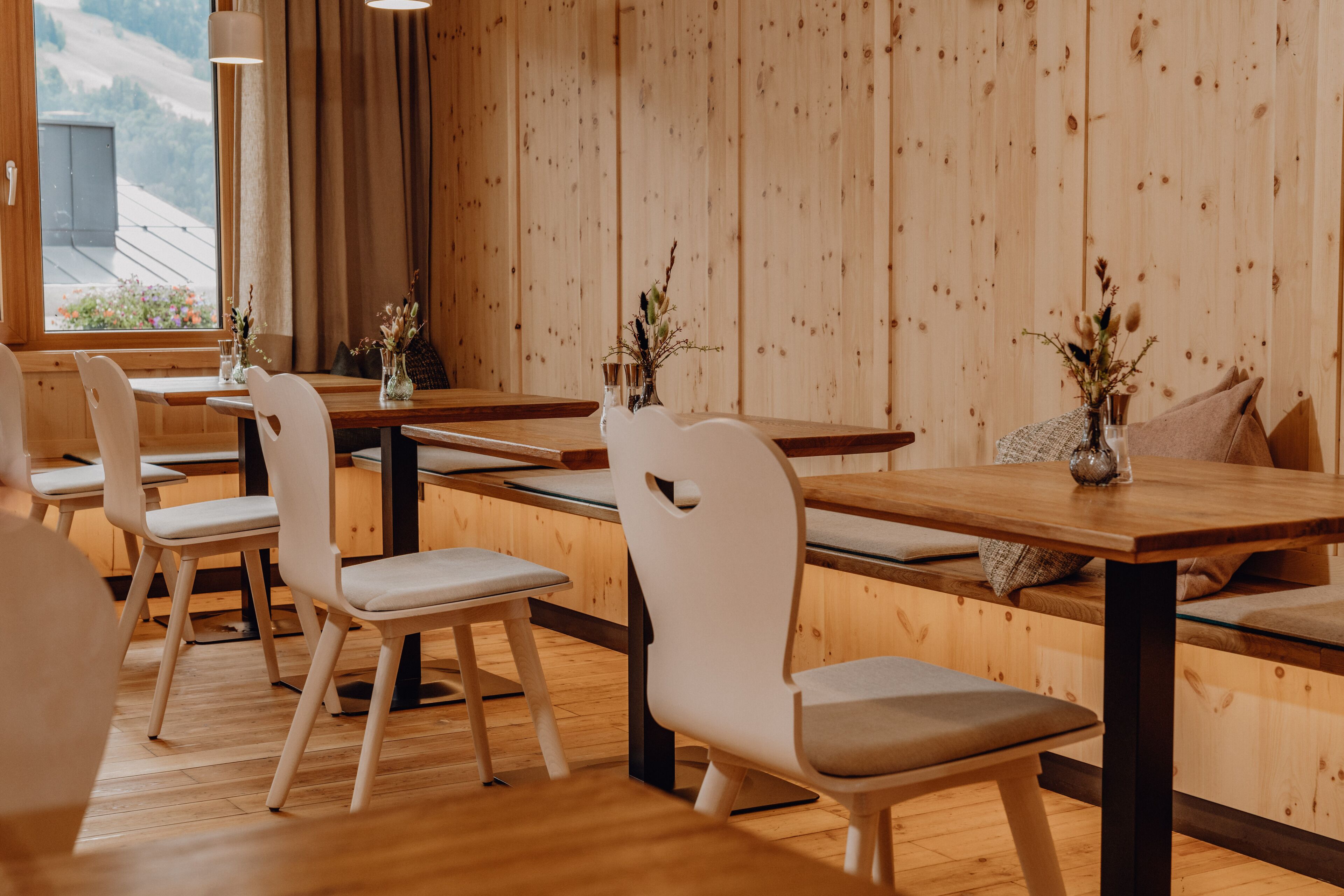 A restaurant with wooden walls and tables, white chairs, and small vases with flowers.