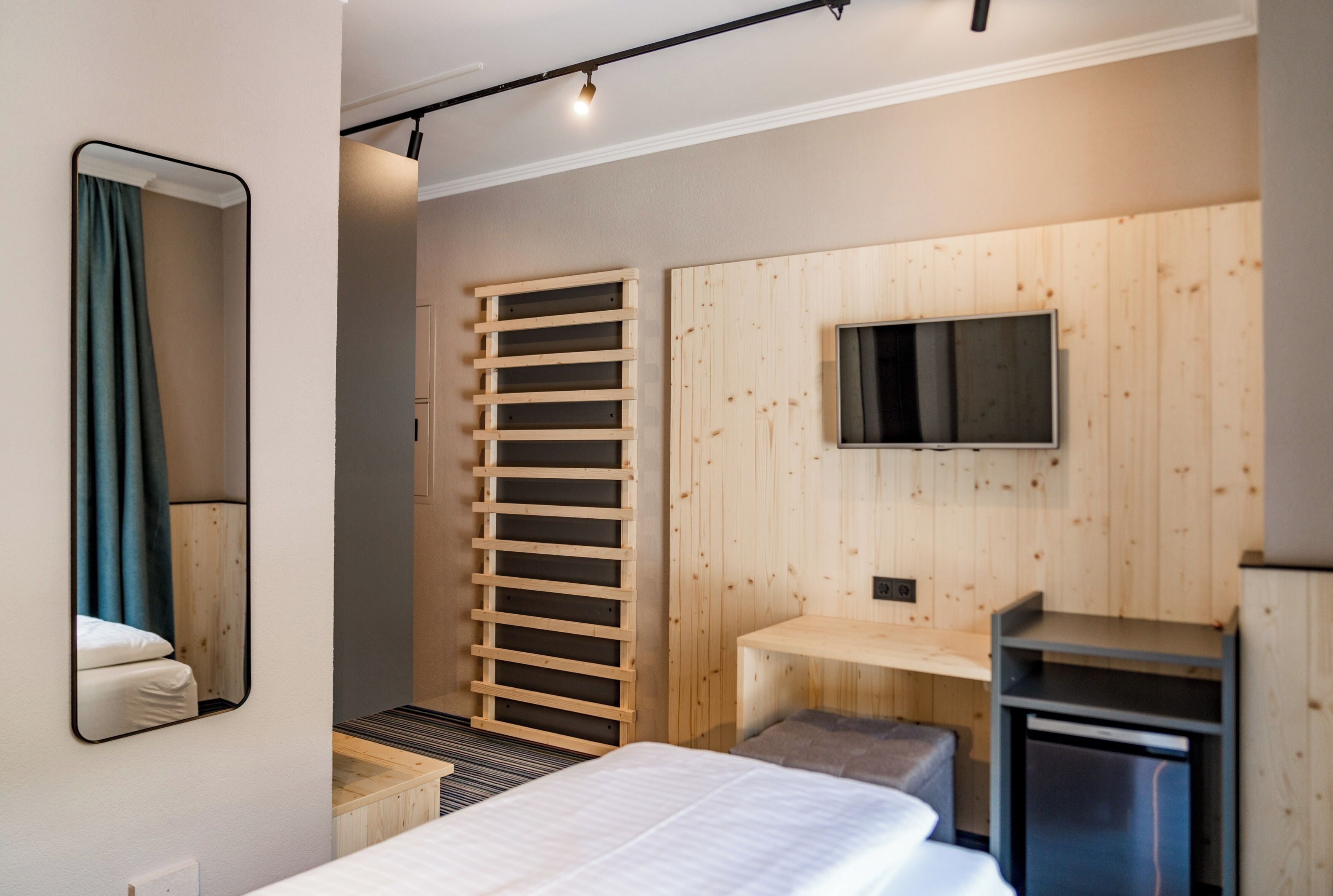 A modern bedroom with a bed, a mirror, a TV mounted on a wooden wall, and a small desk.